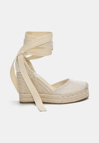 Beiger Keil-Espadrille-Schuh mit geflochtener Seilsohle und langen Stoff-Schnürsenkeln am Knöchel auf weißem Hintergrund.