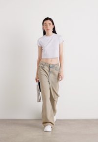 Model loopt in een lichte crop top en oversized beige broek, met een kleine tas in de hand, tegen een neutrale achtergrond. Elegante en casual look.