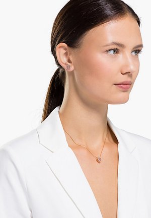 Rose gold jewelry featuring a pendant and stud earrings, intricate design, smooth texture, worn with a white blazer.