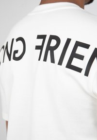 Camiseta blanca con letras negras en negrita que dicen "AMIGO" en la parte posterior, con una textura suave de algodón y mangas cortas.