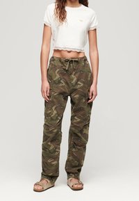 Skåret hvid t-shirt med blonderkant, kombineret med camouflagemønstret cargobukser med sidelommer. Fodtøjet er lysebrune sandaler.