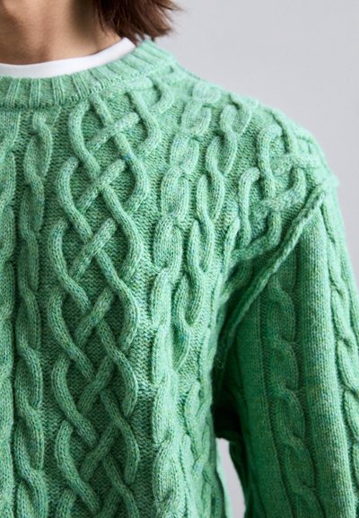 Primer plano de un suéter de punto trenzado verde usado sobre una camiseta blanca en una persona con cabello corto castaño.