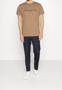 Hnědé bavlněné tričko s grafickým potiskem "TOMMY HILFIGER EST. 1985", doplněné tmavě modrými kalhotami a bílými teniskami. Jednoduchý design a volný střih.