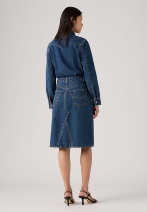Frau steht mit dem Rücken zur Kamera, trägt eine blaue Jeansjacke und einen passenden knieumspielenden Jeansrock mit schwarzen Sandalen mit Absatz.