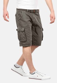 Cargo-Shorts aus olivgrünem Baumwollstoff, ausgestattet mit mehreren Seitentaschen, in lässigem Schnitt und mit einem Gürtel. Kombiniert mit weißen Freizeitsneakern.