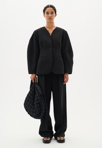Žena stojící v oversize černém blejzru a širokých černých kalhotách, držící velkou prošívanou černou tašku na bílém pozadí.