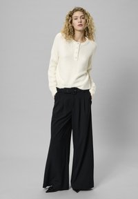 Cremfarbener Strickpullover mit rundem Halsausschnitt und fünf Knöpfen, kombiniert mit hochgeschnittenen schwarzen Hosen mit weitem Bein und Gürtel.