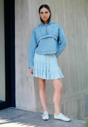 Junge Frau steht drinnen und trägt eine hellblaue, kurze Jeansjacke, einen blassen, gestuften Rock und weiße Turnschuhe vor einem Betonwand-Hintergrund.