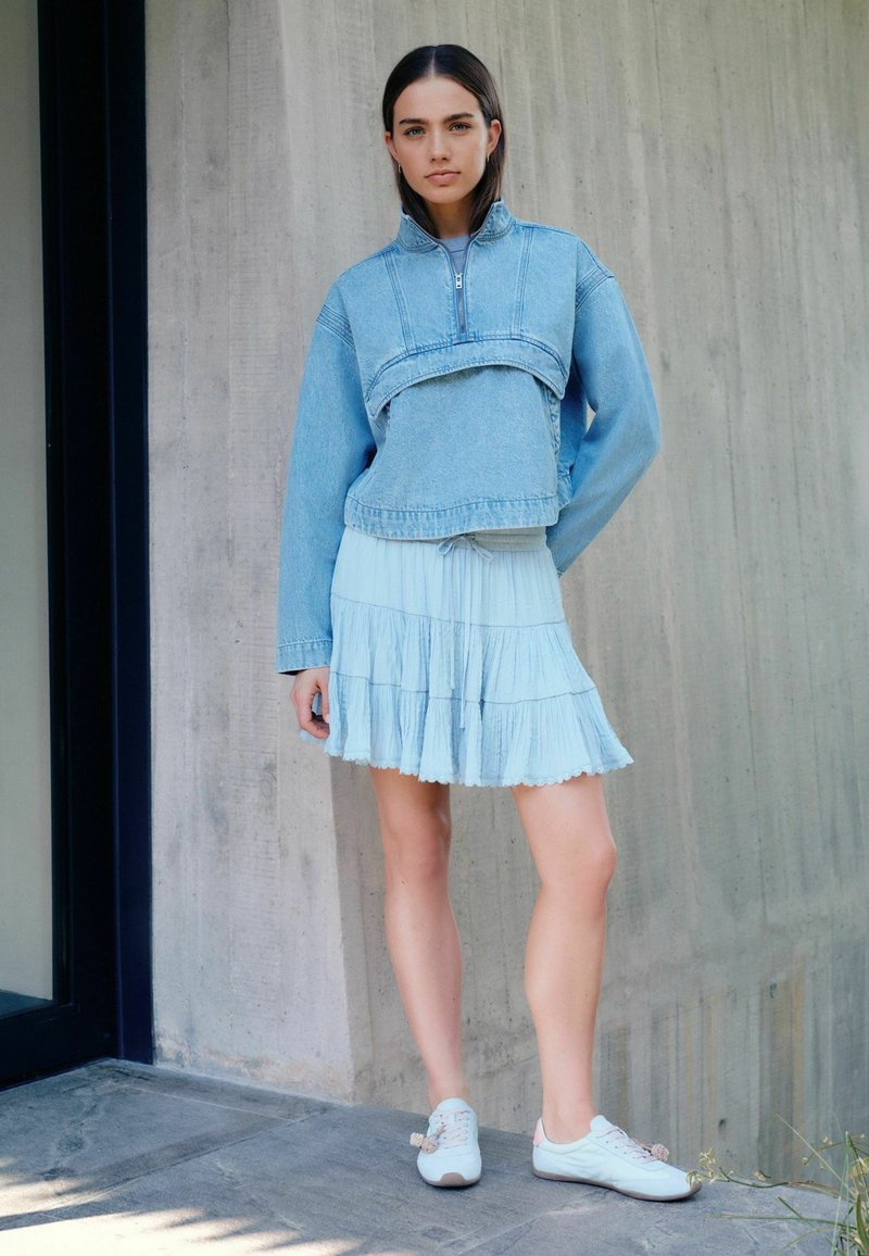 Jeune femme debout à l'intérieur portant une veste en jean courte bleu clair, une jupe claire à plusieurs volants et des baskets blanches, devant un mur en béton.