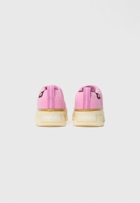 Un par de zapatillas rosas con suelas beige y la marca "SOREL" en el talón, mostradas desde atrás sobre un fondo blanco.