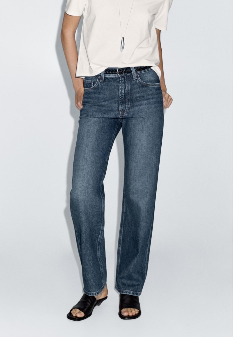 Blå vida jeans med hög midja och femfickorsdesign, kombinerat med en enkel vit T-shirt och svarta flip-flop-sandaler.