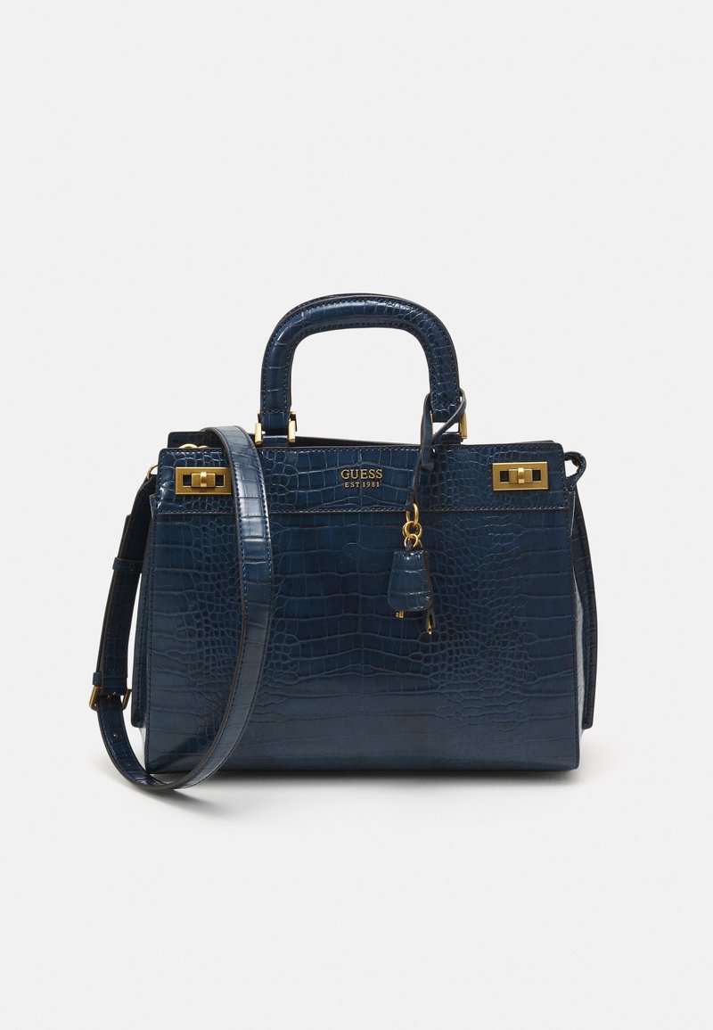 Sac à main bleu foncé à effet crocodile avec finitions dorées, poignées supérieures, bandoulière et logo GUESS sur le devant, sur fond uni.