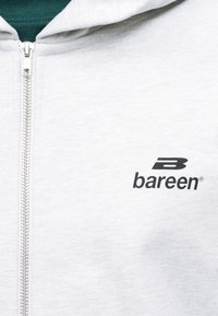 Κοντινό πλάνο ενός ανοιχτό γκρι φούτερ με φερμουάρ σε ασημένιο χρώμα και το μαύρο λογότυπο "bareen" στο στήθος.