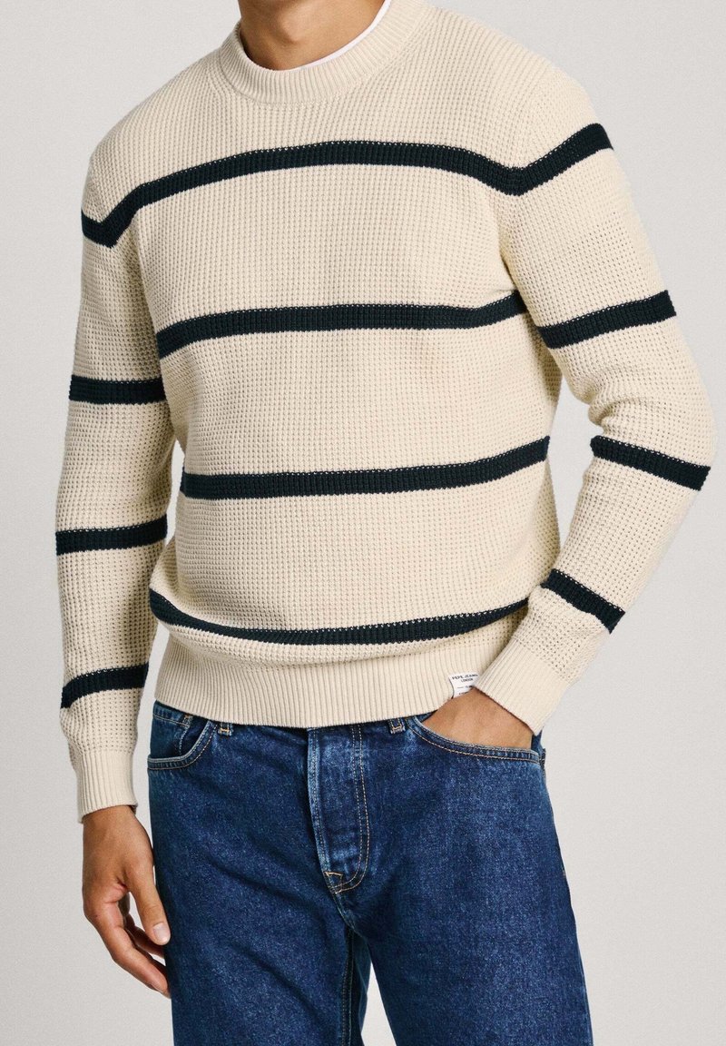 Homme portant un pull en tricot crème avec des rayures horizontales noires et un jean bleu, une main dans la poche, sur un fond uni.