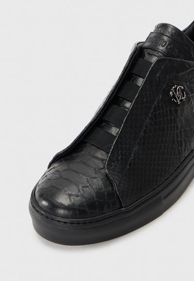 Adidași negri slip-on cu un model texturat de reptilă, benzi elastice în loc de șireturi și un logo metalic al designerului pe lateral.