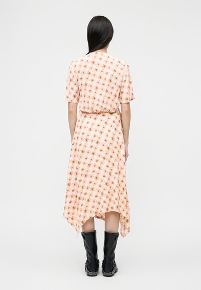 Γυναίκα με μακριά σκούρα μαλλιά φοράει ροζ φόρεμα με λουλούδια και ασύμμετρο τελείωμα, καθώς και μαύρες μπότες μέχρι τη μέση της γάμπας, στέκεται γυρισμένη με την πλάτη σε λευκό φόντο.