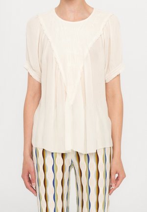 Jasno beżowa bluzka z trójkątną przednią panelą, plisowaną fakturą i krótkimi rękawami. Materiał wydaje się lekki i przewiewny.