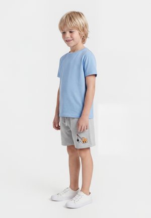 Junger Junge trägt ein hellblaues T-Shirt, graue Shorts mit einem Baukran-Design und weiße Turnschuhe, steht vor einem schlichten Hintergrund.