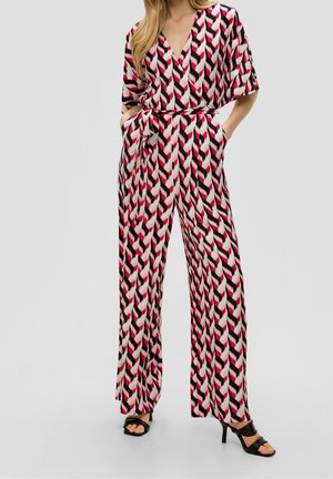 Vrouw draagt een jumpsuit met geometrisch patroon in rood, wit en zwart, met een ceintuur in de taille en wijde pijpen, gecombineerd met zwarte sandalen met hak.