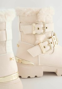 Primo piano di stivali invernali beige eleganti con bordo in pelliccia, fibbie decorative e un dettaglio in oro lucido, su uno sfondo chiaro.