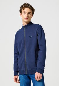 Marinblå zip-up-tröja i mjukt tyg. Har hög krage, ribbade muddar och en liten logotyp på vänster bröst.