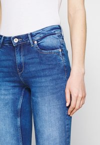 Jeans de mezclilla azul con una cintura de altura media, bolsillos delanteros y una sutil textura desgastada. El diseño incluye un cierre con botón de metal.