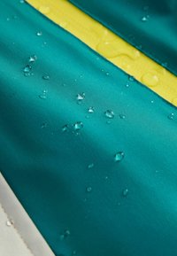 Atsparus vandeniui žalsvai mėlynas audinys su lašeliais, geltona juosta ir lygiu paviršiumi; turi kraštelį su matomu siuvimu.