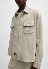 Chemise texturée beige avec un devant à boutons, deux poches poitrine et une étiquette sur la poche gauche. Design à col avec une coupe décontractée.