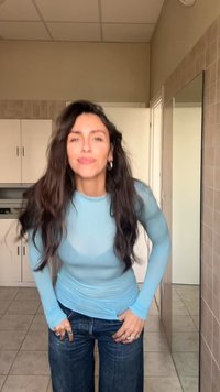 Haut bleu clair à manches longues, semi-transparent, associé à un jean large bleu foncé. Le décor comprend des murs carrelés et des miroirs.