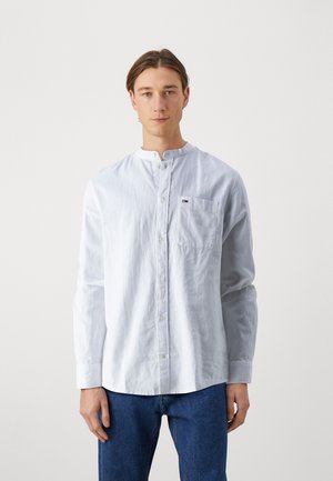 Joven vestido con una camisa blanca de botones con cuello mandarín y jeans azules, de pie contra un fondo claro y liso.