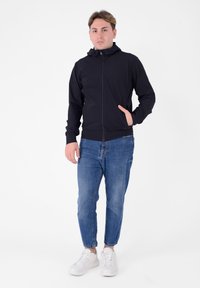 Giacca nera con cappuccio, chiusura con zip, design aderente e polsini elasticizzati, abbinata a jeans azzurri e sneakers bianche.