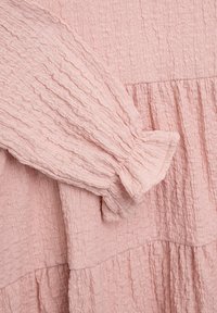 Helles rosafarbenes Kleidungsstück aus strukturiertem, zerknittertem Stoff. Besitzt einen langen Ärmel mit einem gerafften Bündchen und einem schichtweisen Design.