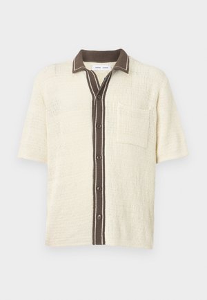 Maglietta a maniche corte in maglia color crema con pattina dei bottoni, collo e dettaglio della tasca sul petto in marrone scuro.