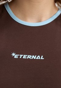 Brun bomullströja med en ljusblå ribbad halsringning. Har en vit logotyp med texten "ETERNAL" samt en stjärn grafisk design.