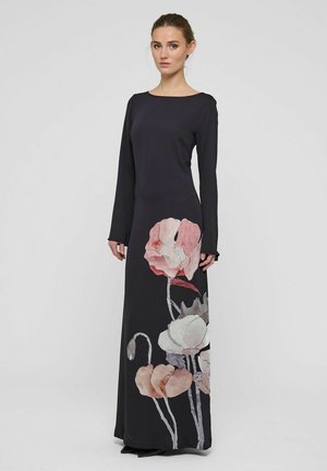 Donna che indossa un lungo vestito nero con stampe floreali rosa e bianche, in piedi davanti a uno sfondo chiaro e uniforme, rivolta in avanti con le mani rilassate.