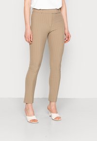 Pantaloni beige a righe sottili con una vestibilità slim e parte anteriore piatta, con vita a media altezza. Abbinati a tacchi bianchi aperti.