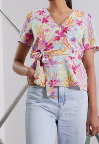 Femme portant un chemisier cache-cœur à imprimé floral rose, orange et violet, associé à un jean bleu clair, tenant un sac vert.