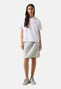 Mujer con el pelo largo y oscuro que lleva una sudadera blanca con capucha de manga corta con diseño rosa, falda gris claro hasta la rodilla y zapatillas beige con detalles rosas.