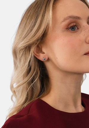 Blonde Frau mit blauen Augen, die ein burgunderrotes Oberteil und kleine silberne, herzförmige Steckerohrringe vor einem einfarbigen Hintergrund trägt.