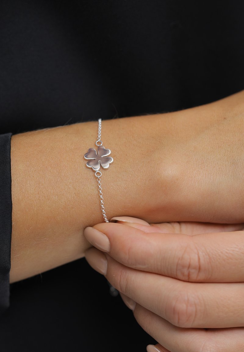 Bracelet en argent avec un pendentif en forme de trèfle, présentant une chaîne délicate et une surface polie. Teint de peau visible, les ongles sont de couleur neutre.