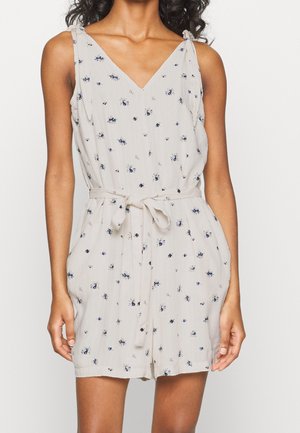 Ærmeløs beige romper med lille blåt blomsterprint, V-udskæring, stofbælte bundet i taljen og løstsiddende shorts.