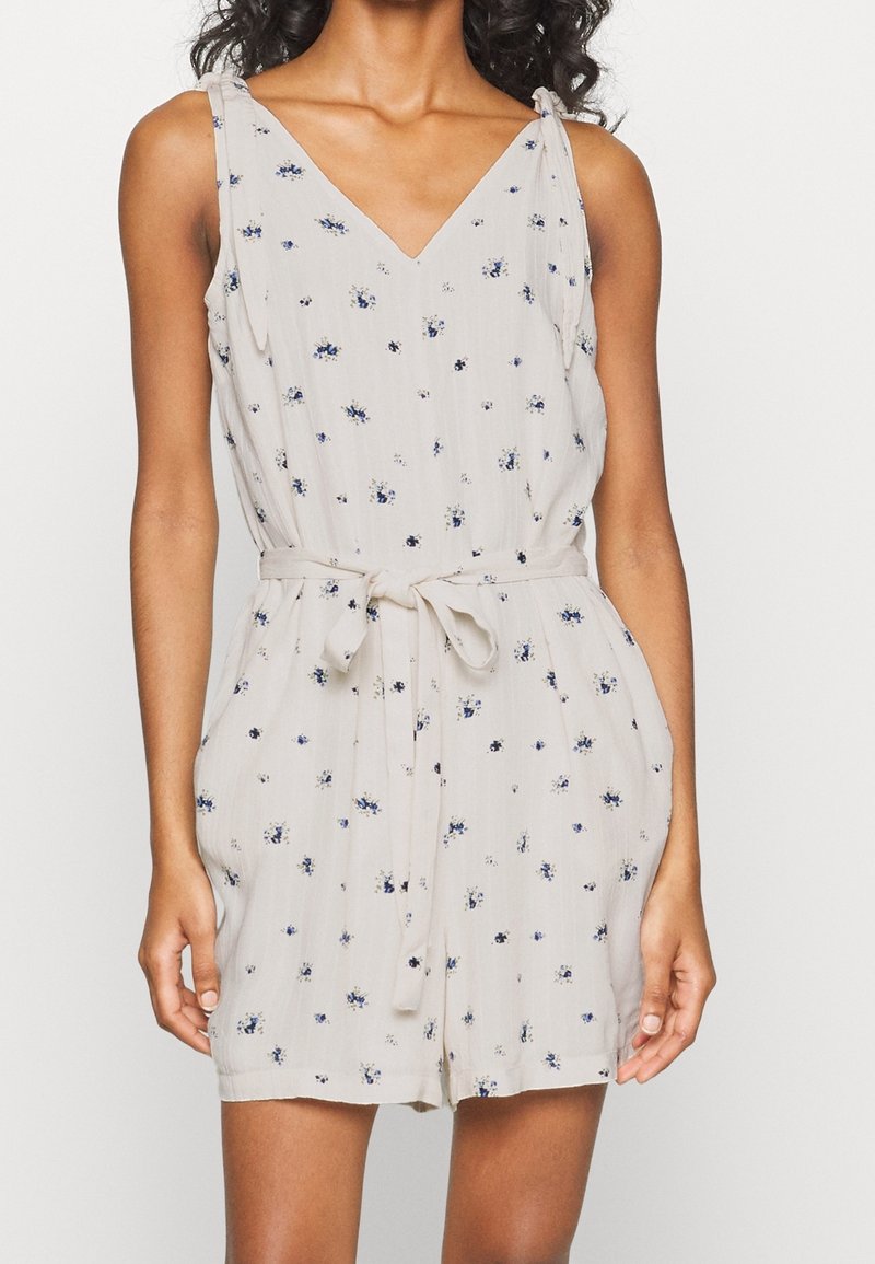 Combinaison sans manches beige avec petit motif floral bleu, encolure en V, ceinture en tissu nouée à la taille, et shorts amples.