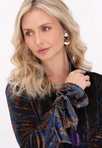Een fluwelen blouse met een marineblauw en roestkleurig paisleypatroon, voorzien van een vastgebonden manchet met vloeiende mouwen. Statement oorbellen met neutrale en gekleurde accenten.