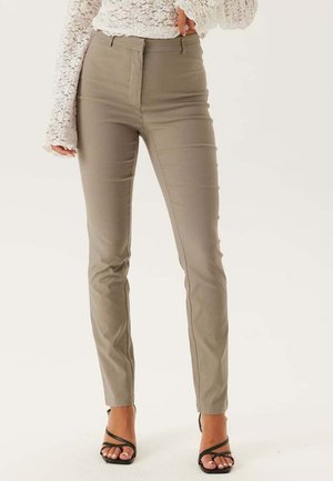 Pantaloni beige a vita alta con vestibilità aderente, texture liscia e dettagli con cucitura centrale, abbinati a sandali neri con cinturini.