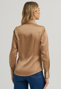 Brązowa satynowa koszula z zaokrąglonym karczkiem, długimi rękawami i zapięciem na guziki. Gładka tekstura i dopasowany krój w połączeniu z niebieskimi dżinsami.