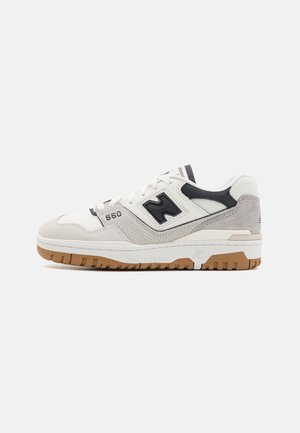 Biało-szary but sportowy New Balance 550 z czarnym logo i gumową podeszwą, widok z boku na jednolitym jasnoszarym tle.