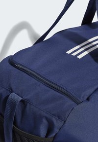 Námornícky modrý batoh vyrobený z odolnej tkaniny, ktorý má zipsovú kapsu, textúrovaný povrch a tri biele pruhy ako pozoruhodný dizajnový detail.