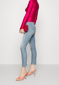 Vaqueros ajustados azul con un acabado desgastado, combinados con una blusa de manga larga rosa vibrante y mules de tacón color coral. Sosteniendo un clutch de cuentas rojas.