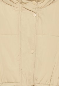 Beige péřová bunda s kapucí, která má patentky a viditelné stehy. Nastavitelná šňůrka na lemu. Hladká, lehká tkanina.