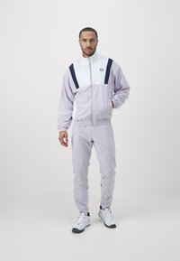 Lila und weiße Trainingsanzug mit elastischen Bündchen und einem elastischen Taillenbund, ausgestattet mit einem Logodetail und kontrastierenden navy Streifen. Getragen mit weißen Schuhen.